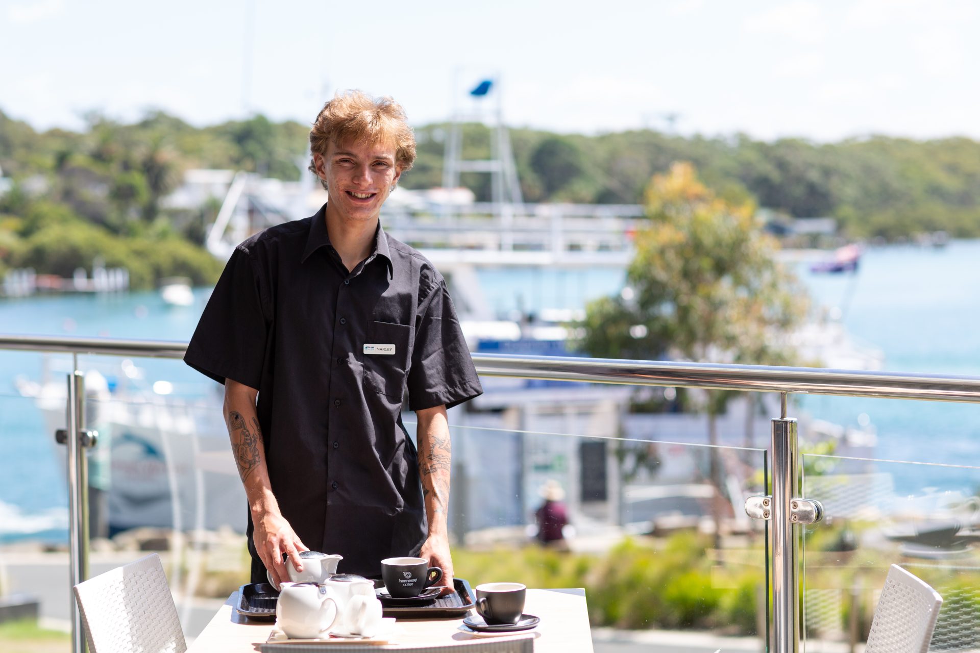 Young man in a black uniform serving coffee on an outdoor deck, whose supported by the personnel group and iea